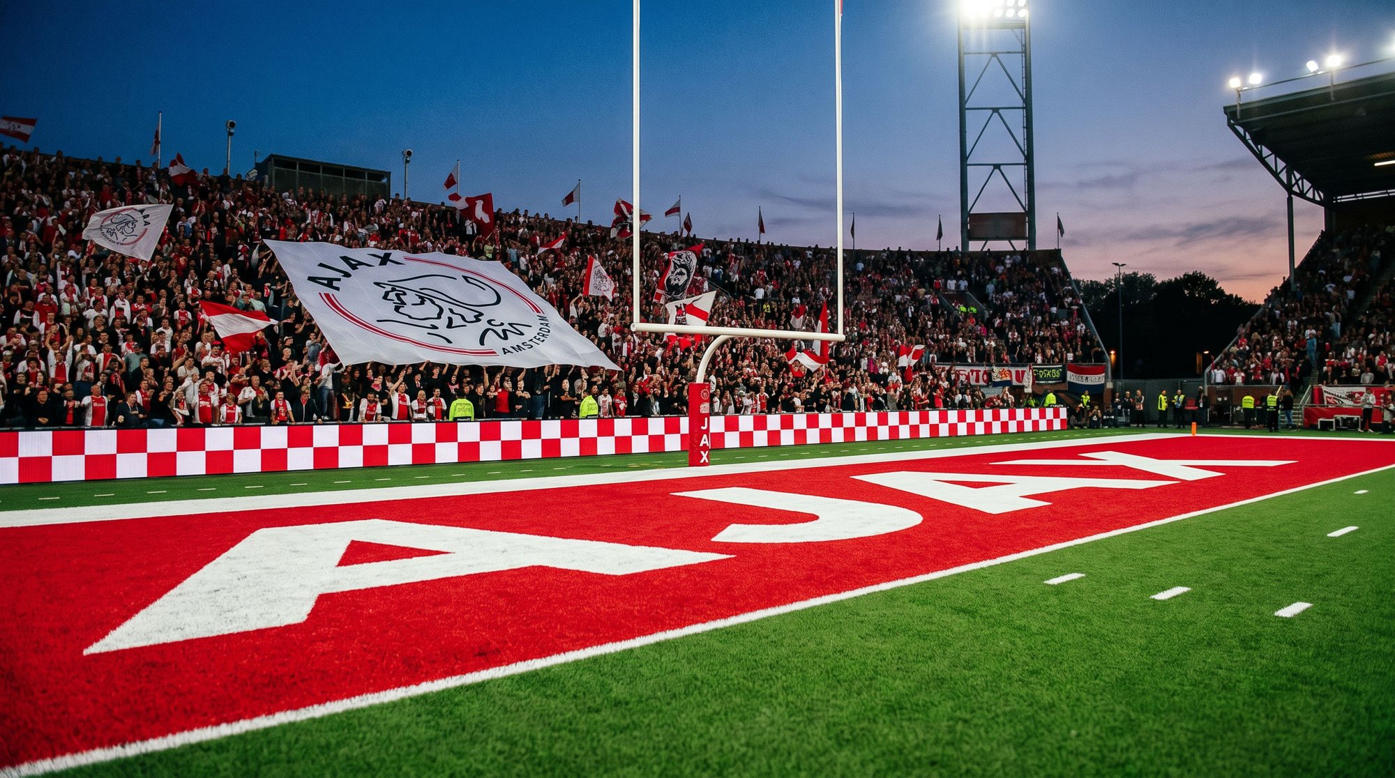 Ground-level view into the red AJAX end zone at dusk, a white goal post and a crest banner in the packed stand