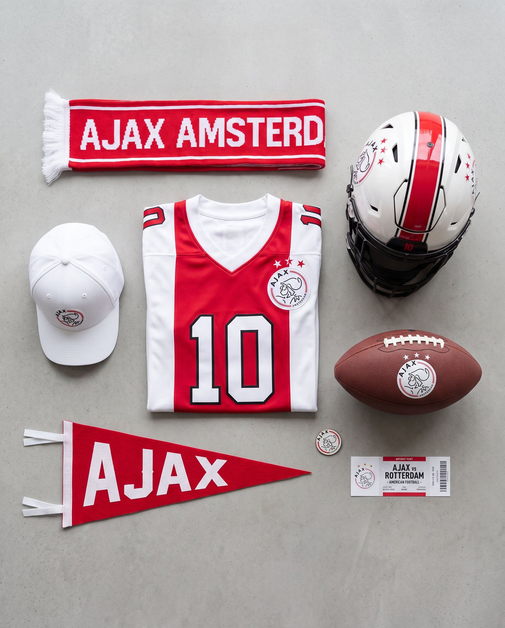 Flat lay of Ajax football merchandise: home jersey 10, mini helmet, AJAX AMSTERDAM scarf, cap, leather football with the crest, pennant, pin and matchday ticket