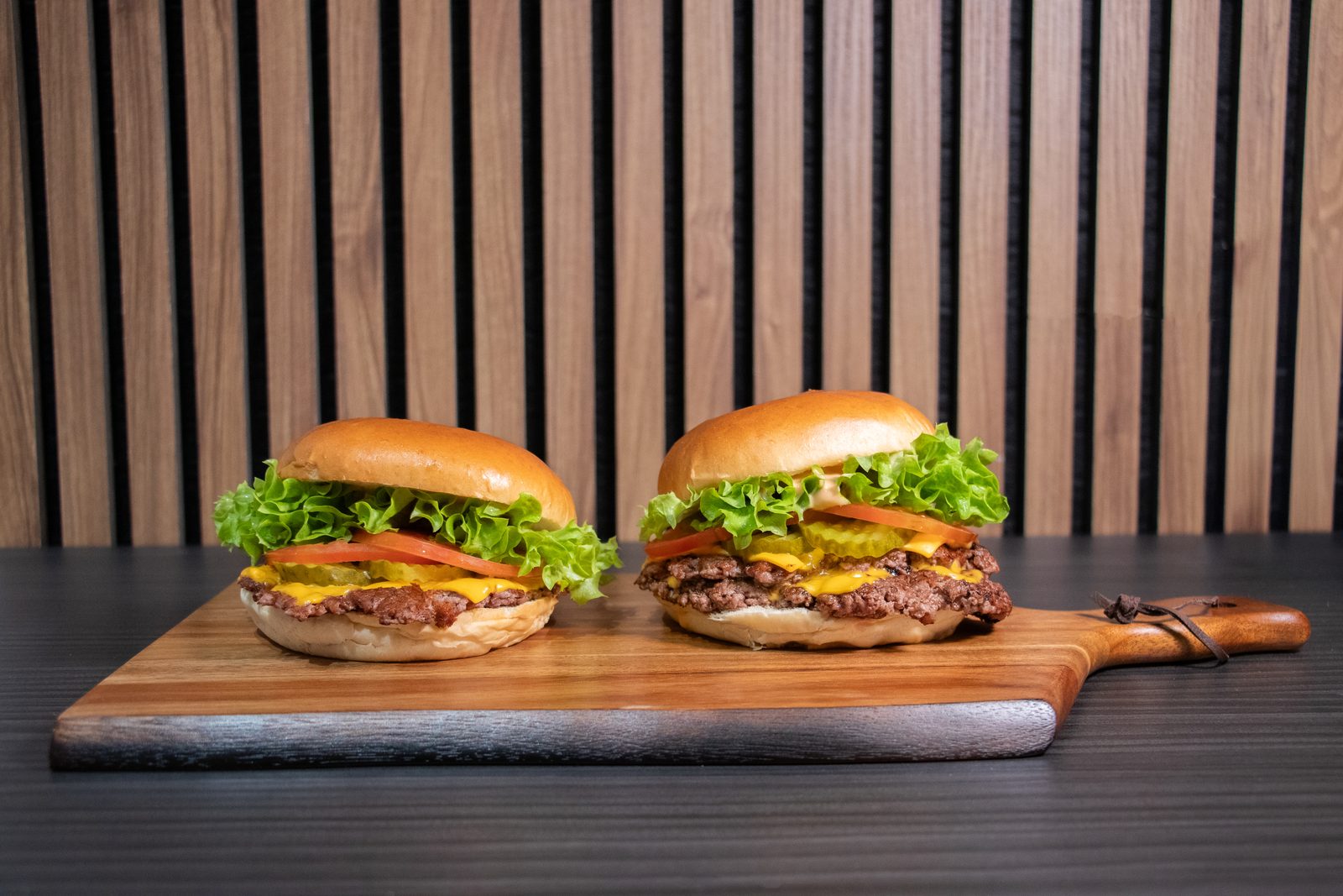 Two burgers on a wooden board in front of a slatted wood wall