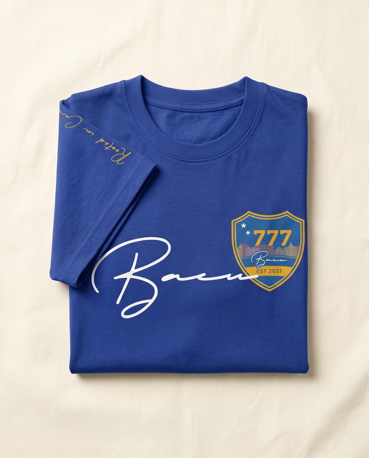 Folded blue BACU tee with script logo and 777 crest on cream fabric