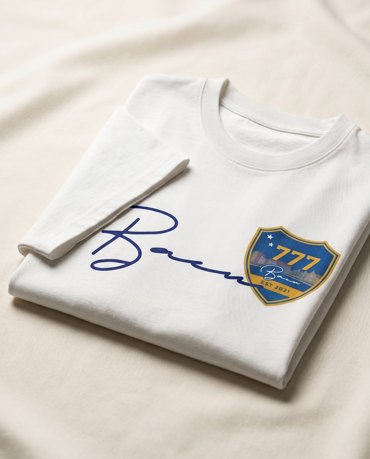 Folded white BACU tee with script logo and crest on cream fabric