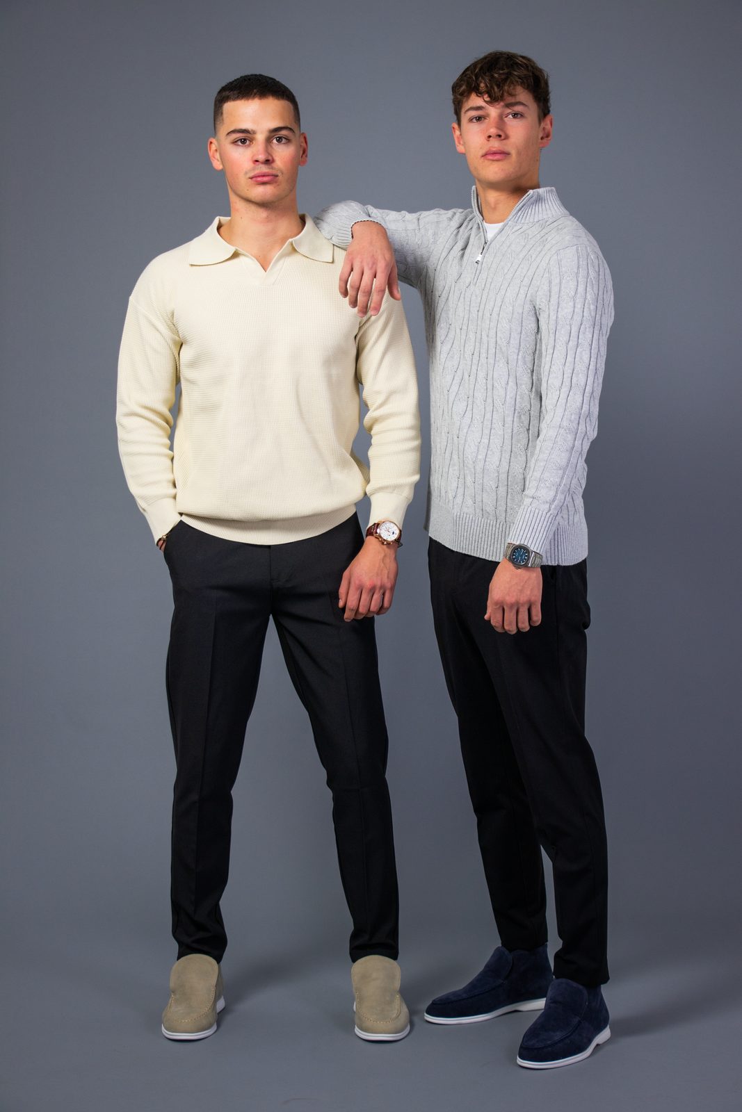 Two models in cream and grey cable-knit NOBA sweaters on a grey backdrop