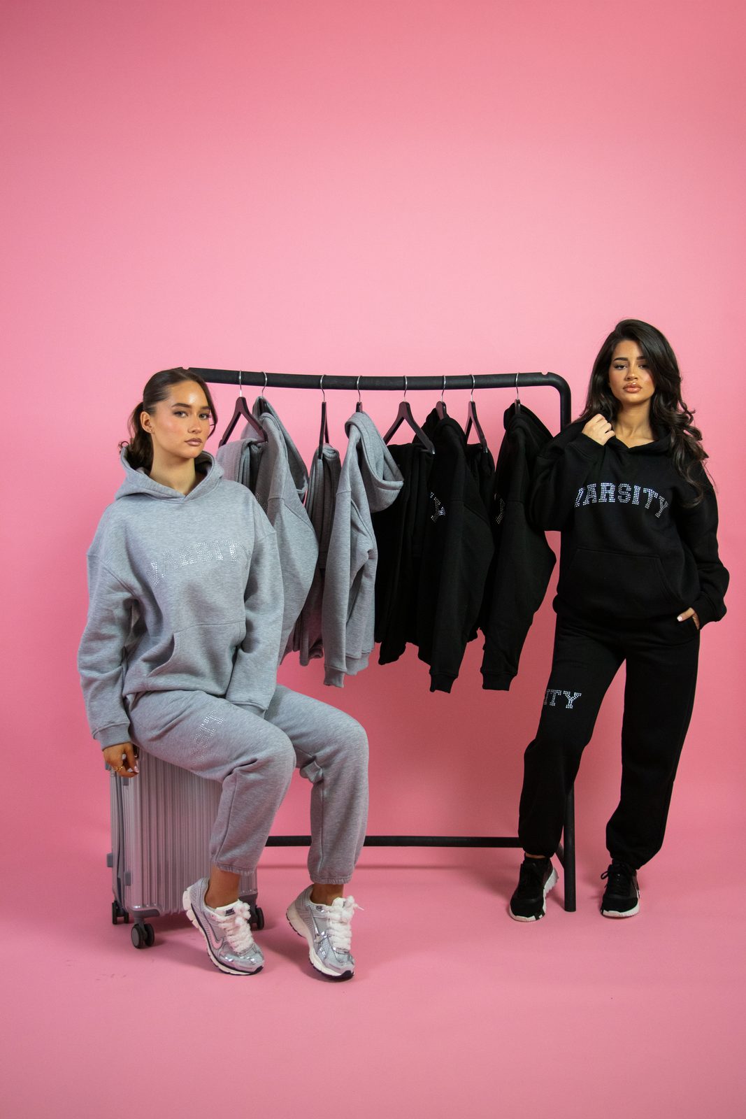 Two models in grey and black Varsity tracksuits by a clothing rail on a pink backdrop