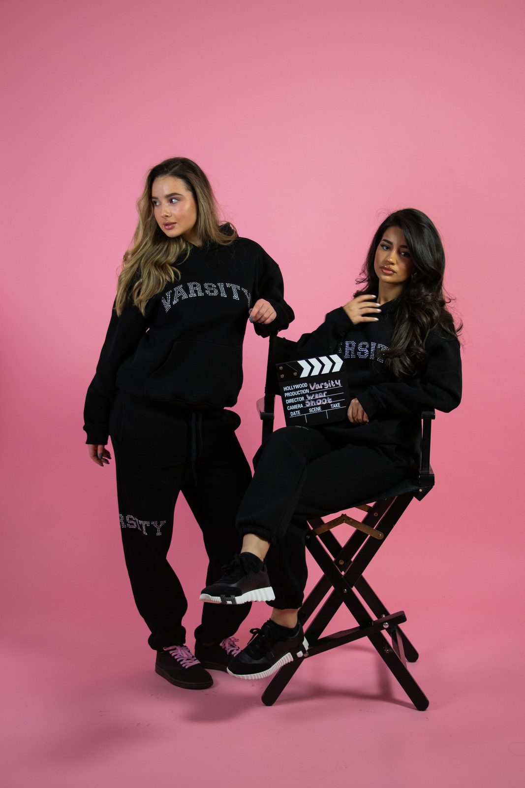 Two models in black Varsity sets with a director's chair on pink