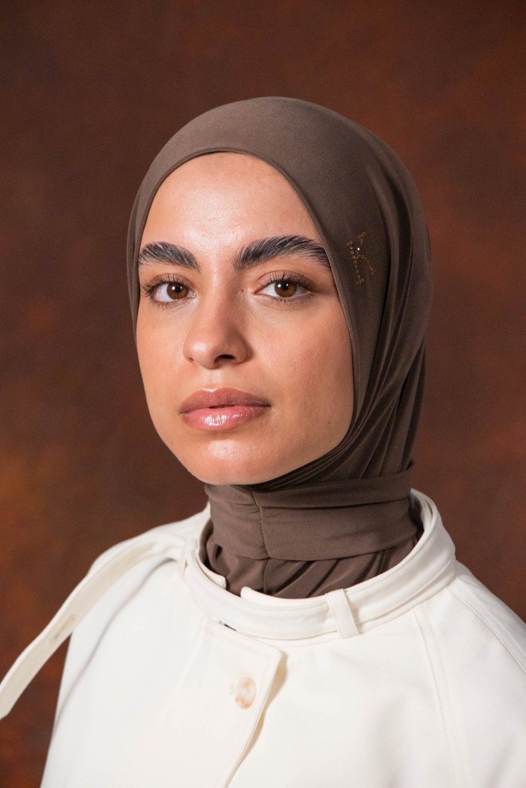 Model in a brown hijab and ivory jacket against a brown backdrop