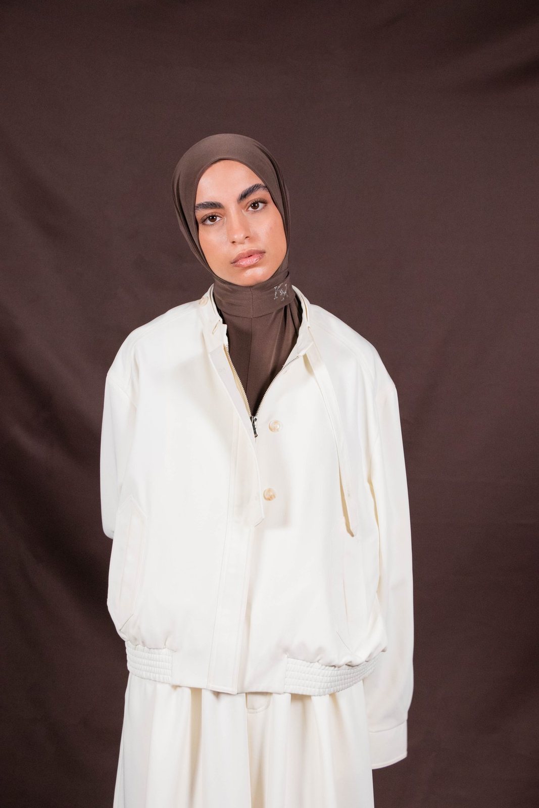 Model in a brown Kallouchi hijab and ivory jacket against a brown studio backdrop