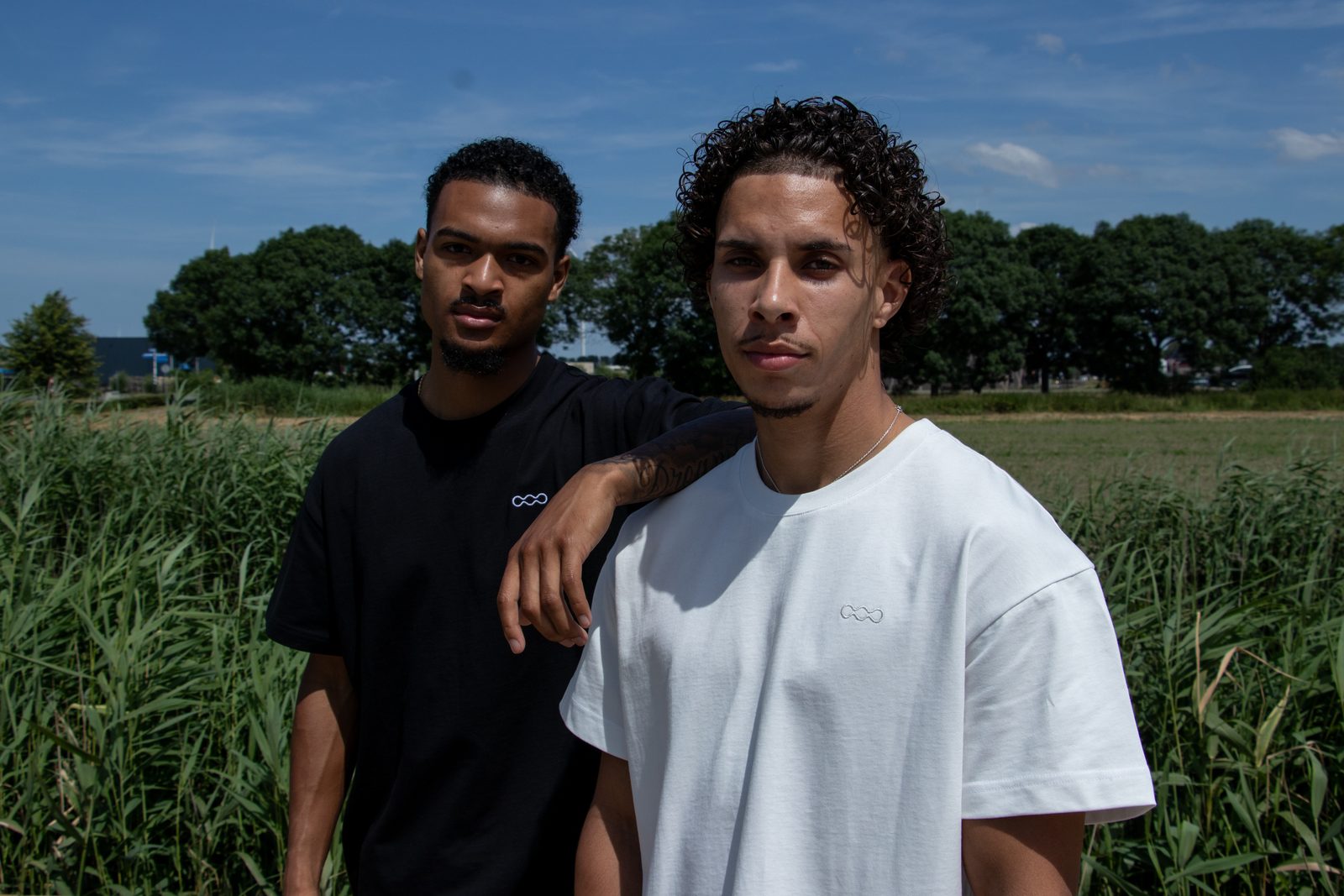 Two models in black and white LMF tees in a field of tall grass under a blue sky