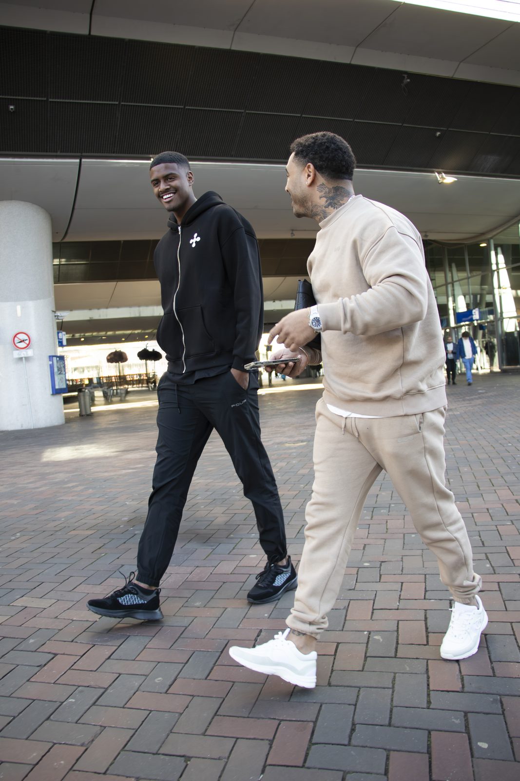 Two models in black and beige LMF tracksuits walking and laughing