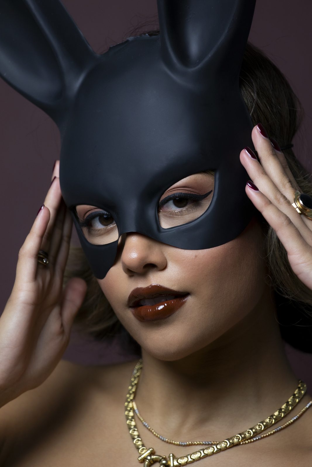 Model in a black bunny mask with dark brown lipstick and gold chains against a deep burgundy backdrop