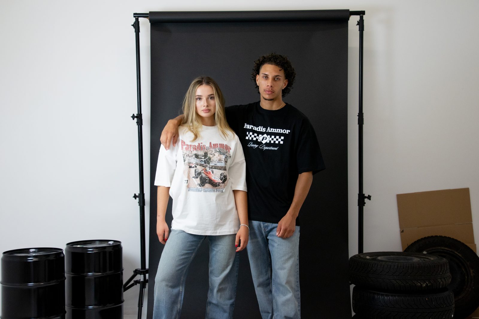 Two models in Paradis Ammor racing tees in front of a black backdrop with tyres and oil barrels