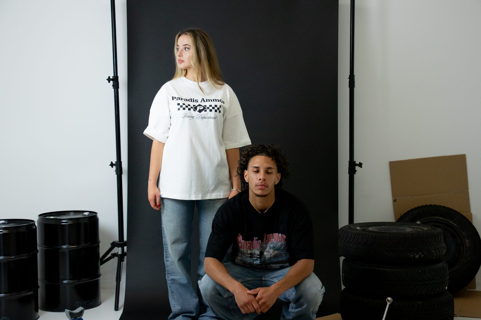 One model standing and one crouching by the tyres in Paradis Ammor tees