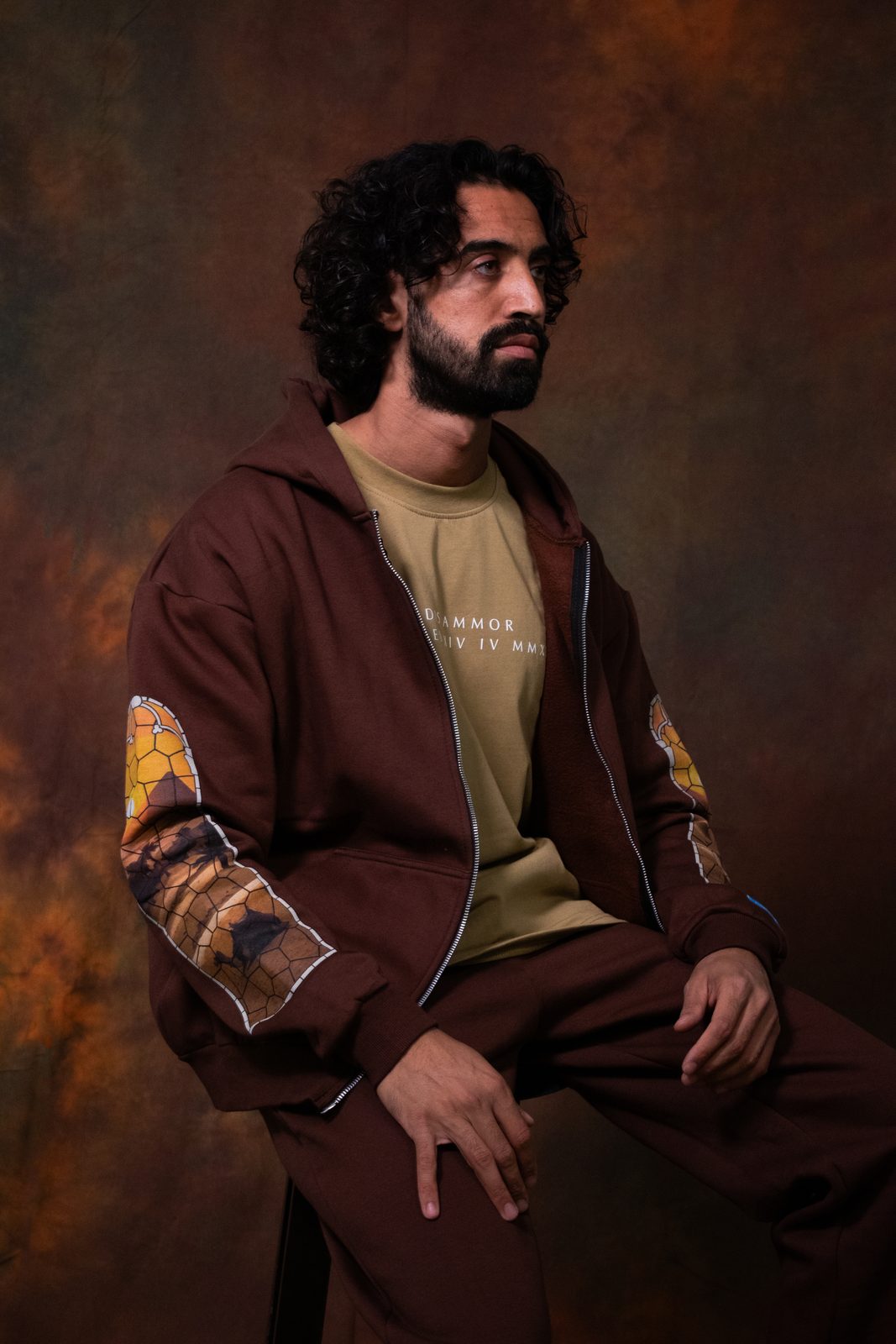 Model seated in a brown zip hoodie with printed sleeves against a painted backdrop