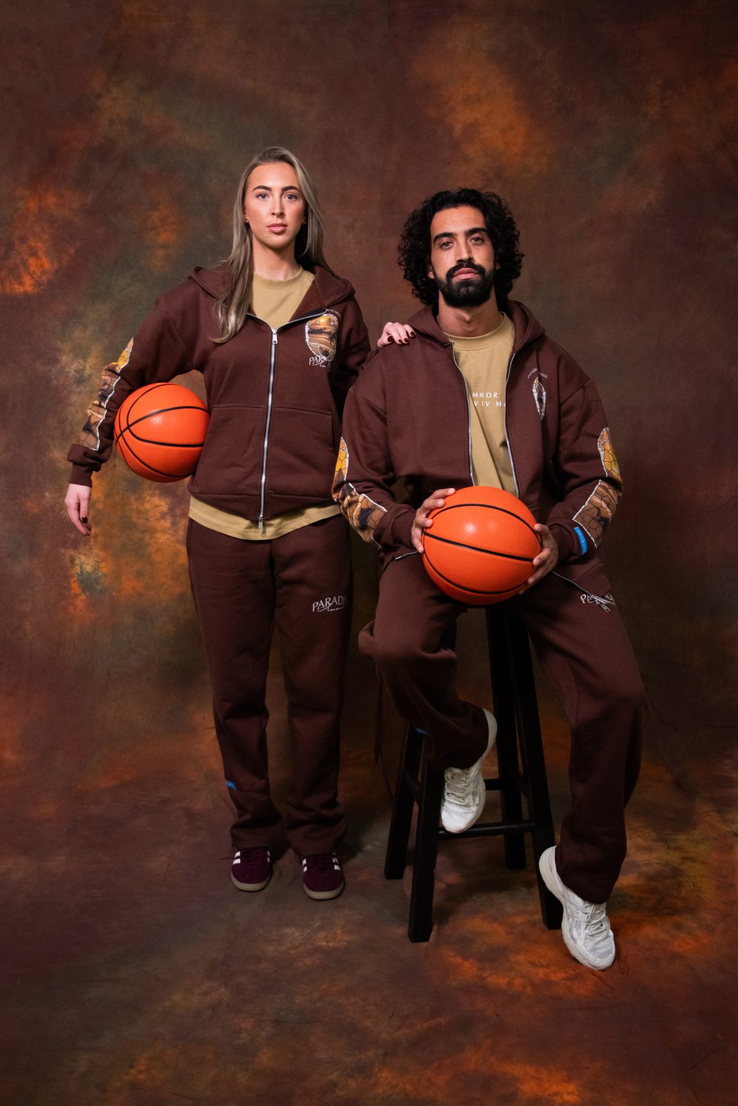 Two models in matching brown Paradis Ammor zip hoodies and trousers holding basketballs on a stool in front of a painted backdrop
