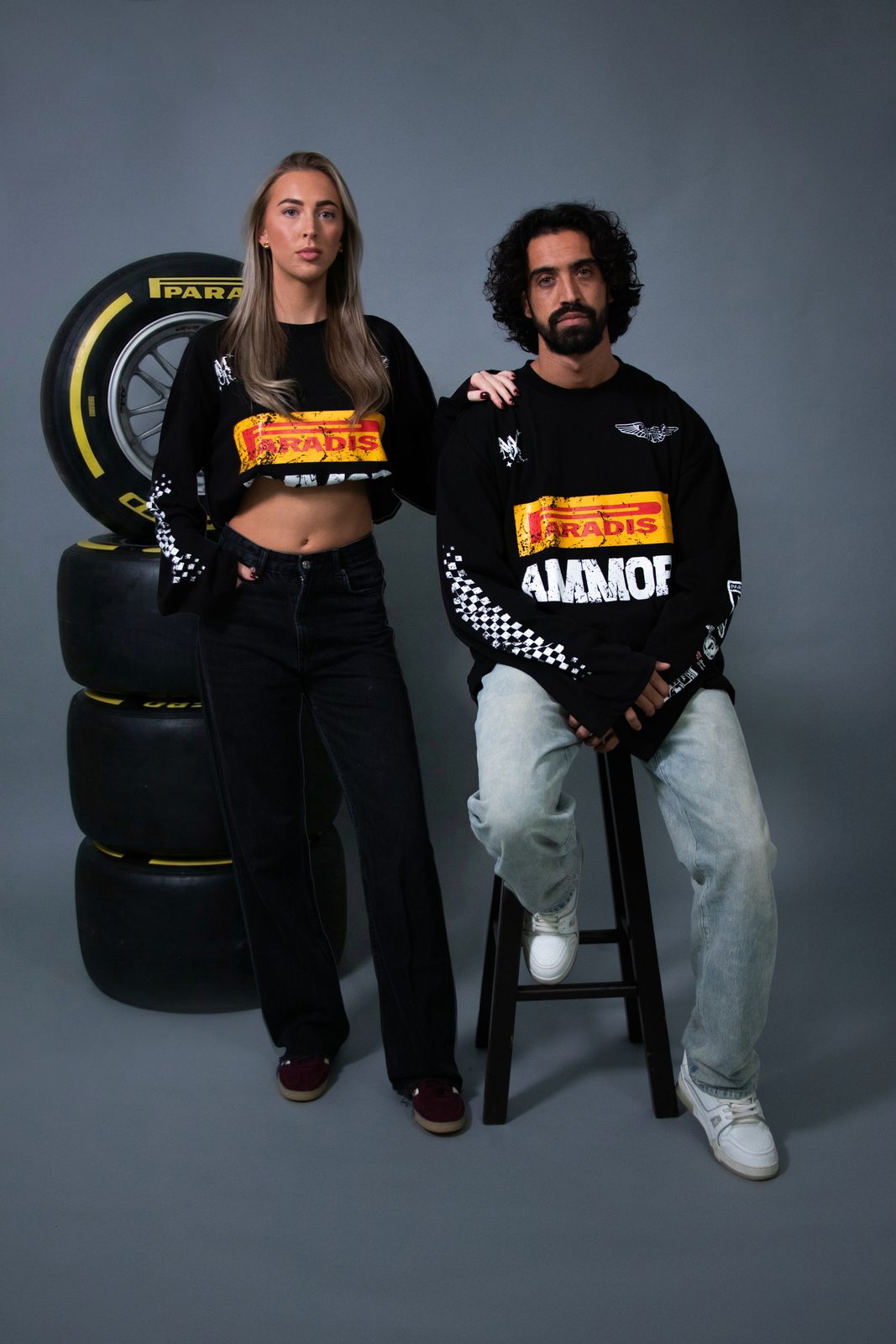 Two models in black racing longsleeves next to a stack of race tyres