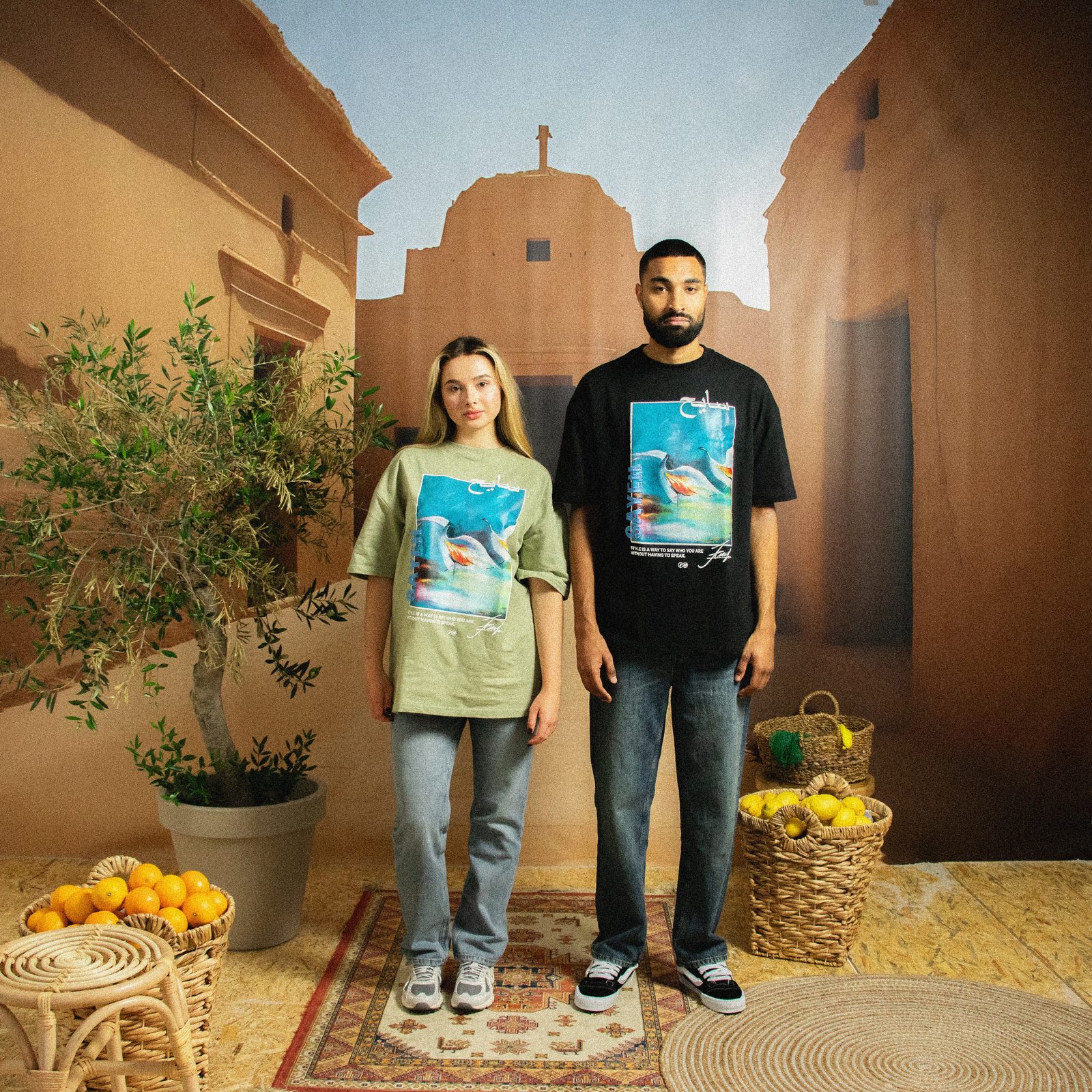 Two models in SAYEH tees on a Marrakech set with a painted courtyard backdrop, an olive tree, rugs and baskets of oranges
