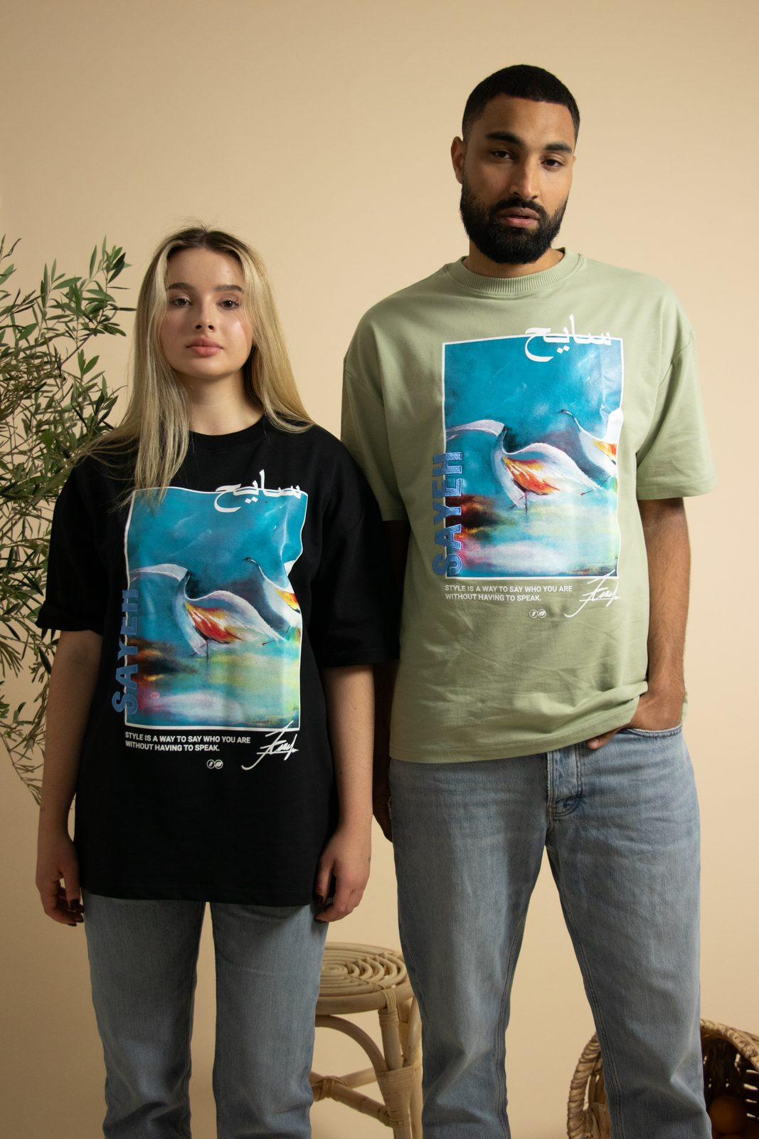 Two models in black and sage SAYEH printed tees against a beige wall