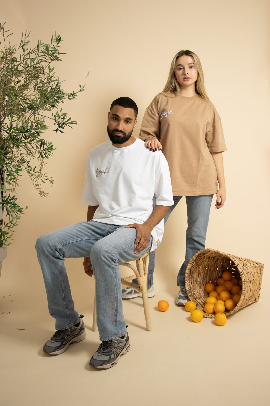 Model seated on a rattan stool with a second model behind him and spilled oranges
