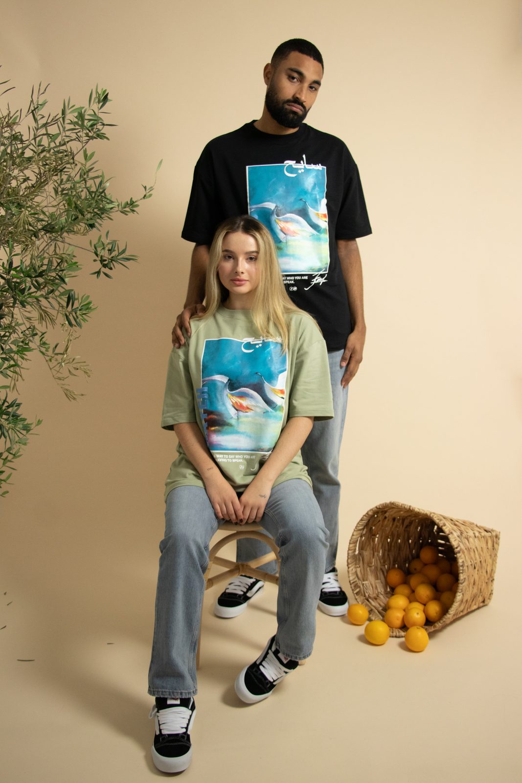 Two models in SAYEH printed tees on a rattan stool next to a basket of spilled oranges