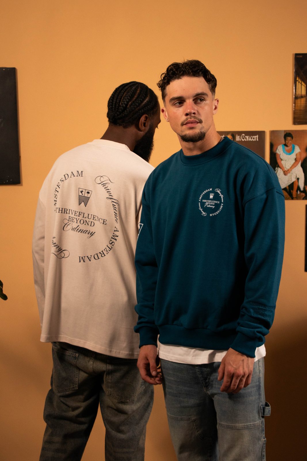 Two models in Thrivefluence sweatshirts on a warm orange living-room set with records on the wall