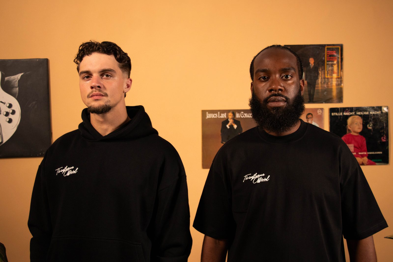 Two models in black Thrivefluence hoodie and tee on an orange living-room set